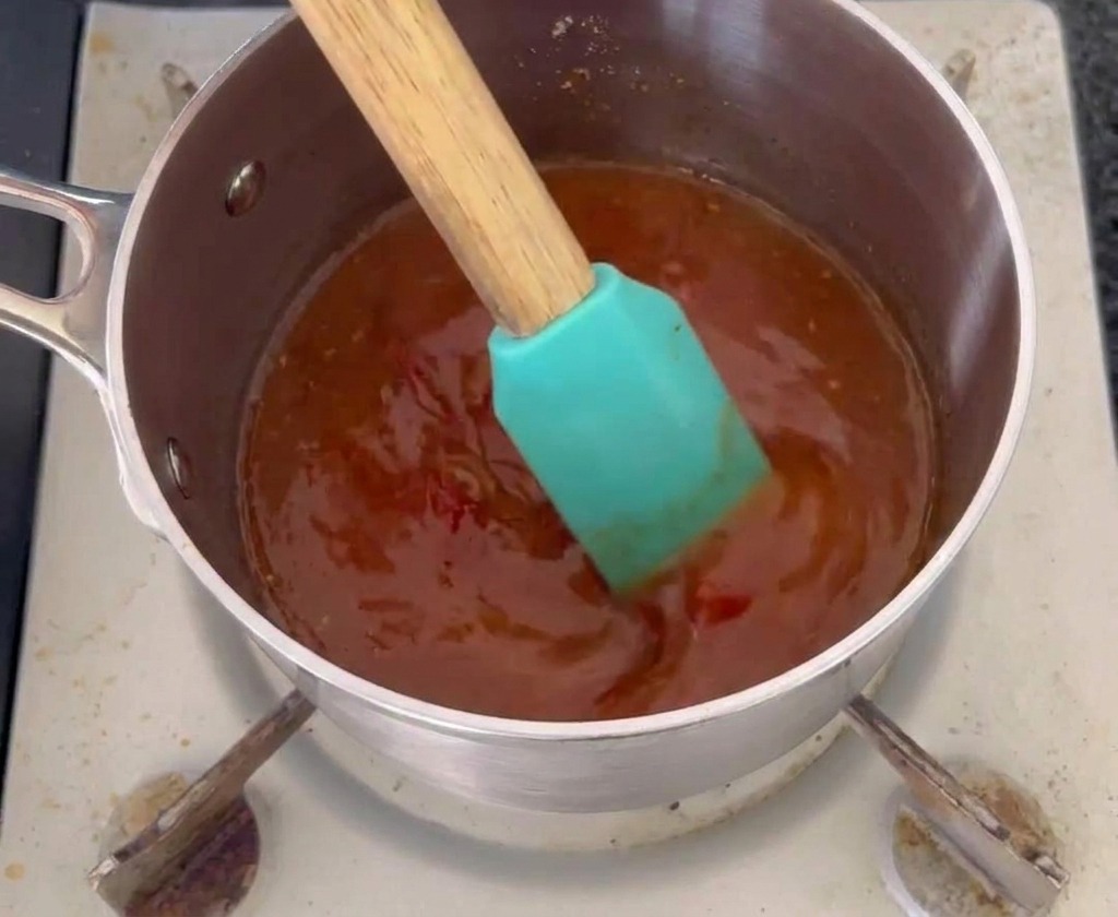 Una espátula de silicona de color turquesa removiendo una salsa mop de color naranja rojizo oscuro en una pequeña olla de acero inoxidable.