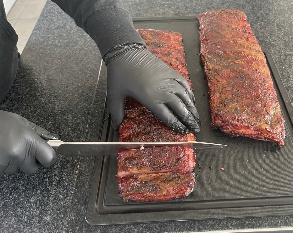 Una persona con guantes de nitrilo negros utilizando un gran cuchillo de chef para cortar un costillar de cerdo ahumado de color rojo oscuro sobre una tabla de cortar negra.