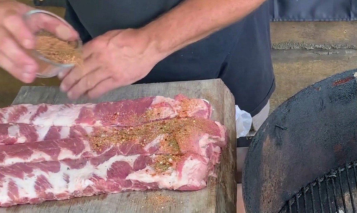 Manos de una persona espolvoreando una mezcla de especias de color marrón rojizo sobre costillas de cerdo crudas en una tabla de picar de madera.
