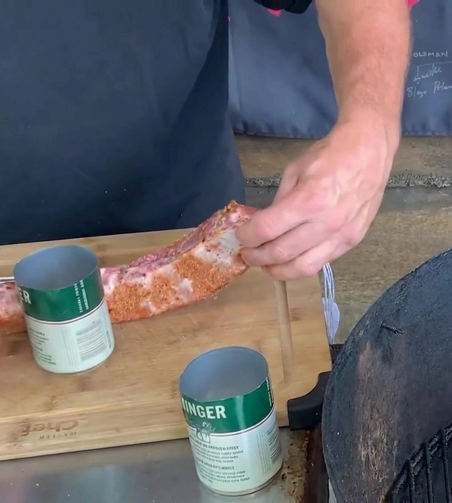 Un par de manos wrapping una costilla de cerdo sazonada alrededor de una lata de cerveza metálica verde y blanca sobre una tabla de cortar.