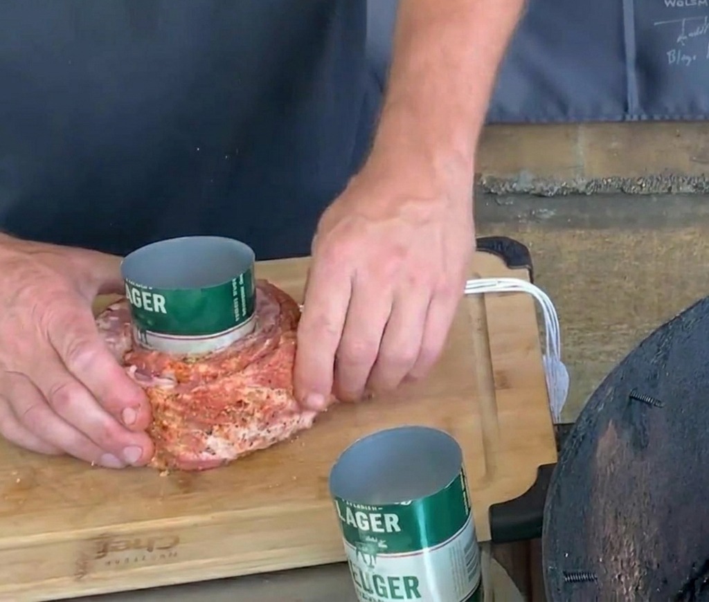 Manos asegurando cuidadosamente hilo de carnicero blanco alrededor de una costilla de cerdo sazonada envuelta en una lata de cerveza verde.