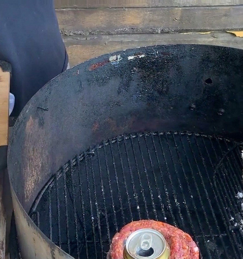 Una copa de costilla amarrada que contiene una lata de cerveza de metal sobre la rejilla de una parrilla de carbón negra al lado de una tabla de preparación.