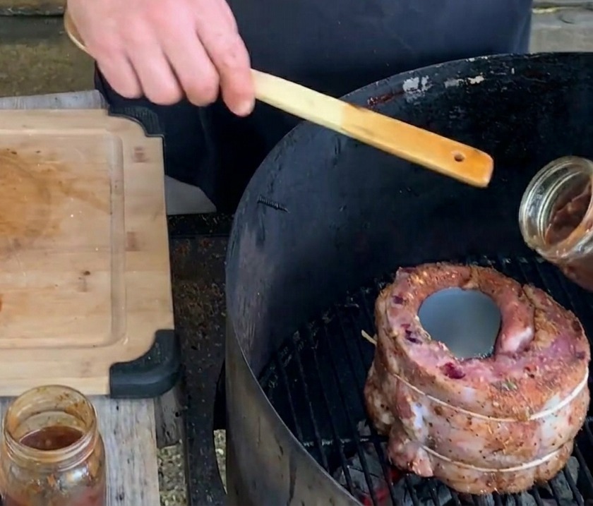 Una mano vierte una salsa espesa y oscura de arándanos desde un frasco de vidrio en una lata de cerveza de metal centrada dentro de una copa de costilla en la parrilla.