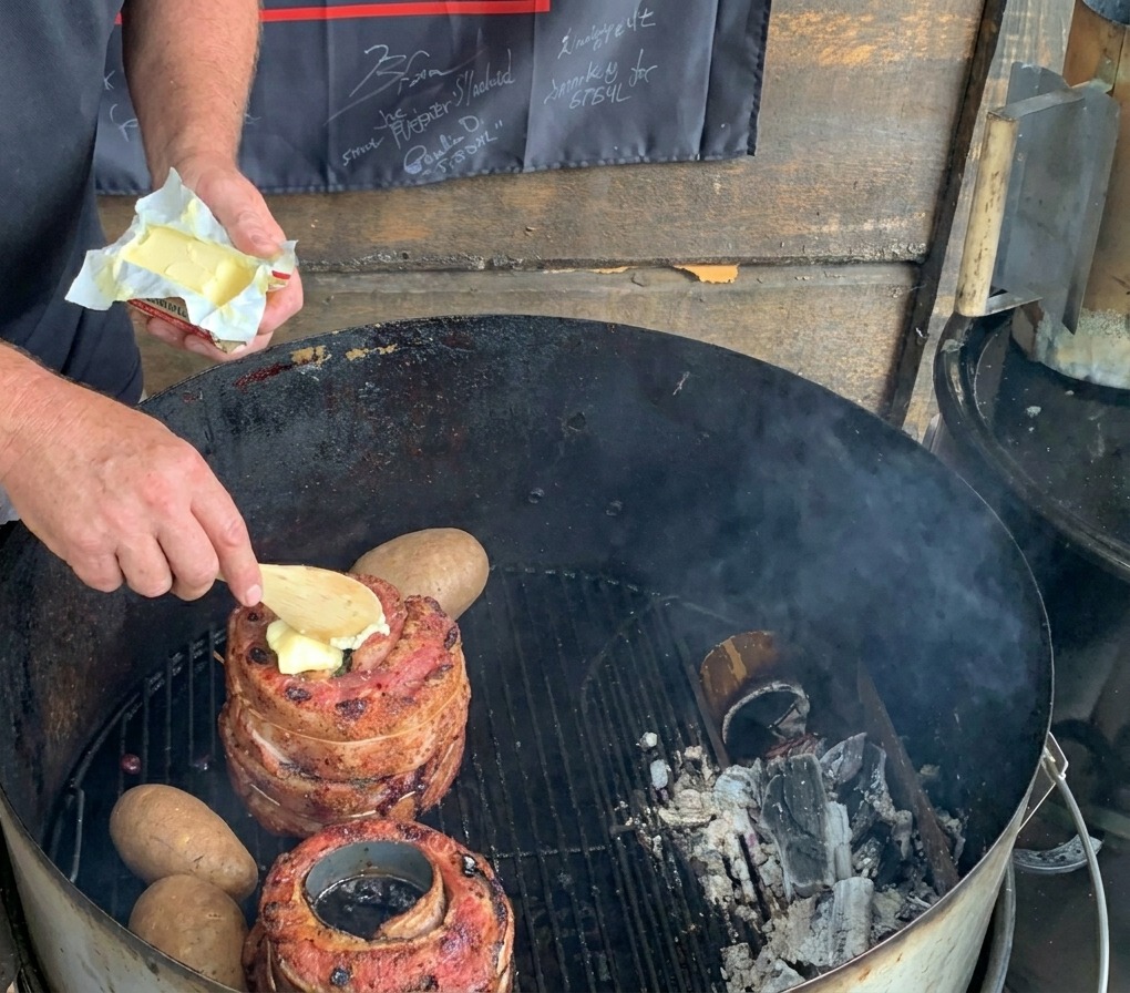 Una cuchara de madera colocando un trozo de mantequilla sobre la carne en una parrilla de carbón junto a papas enteras.
