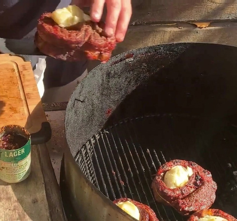 Una persona colocando una copa de costilla rellena sobre la rejilla de la parrilla junto a otras copas de costilla y tiras de tocino.