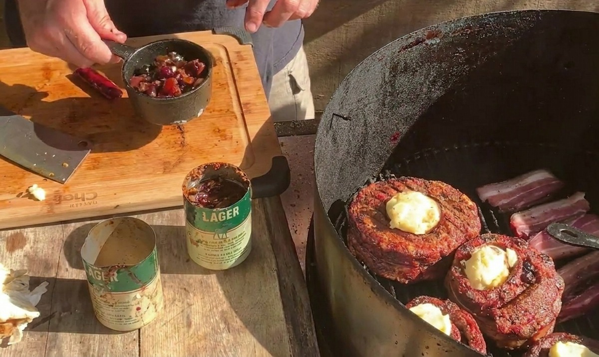 Una persona sosteniendo una pequeña olla de metal llena de salsa de arándanos sobre una parrilla donde las copas de costilla envueltas en tocino se cubren con queso.