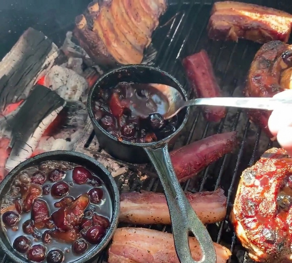 Una persona usando una cuchara para verter salsa oscura de arándanos desde una pequeña olla de hierro fundido sobre copas de costilla a la parrilla rellenas con papas.