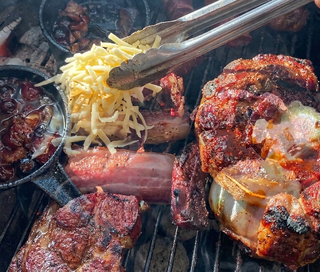 Unas pinzas dejando caer queso amarillo y blanco rallado sobre una copa de carne a la parrilla rellena con una papa y salsa de arándanos.
