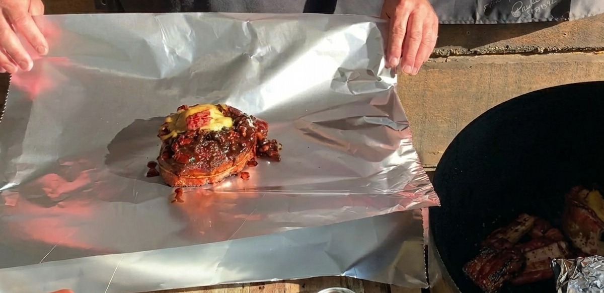 Una copa de costilla BBQ terminada y cubierta con queso fundido reposando sobre una gran hoja de papel de aluminio, lista para ser envuelta.