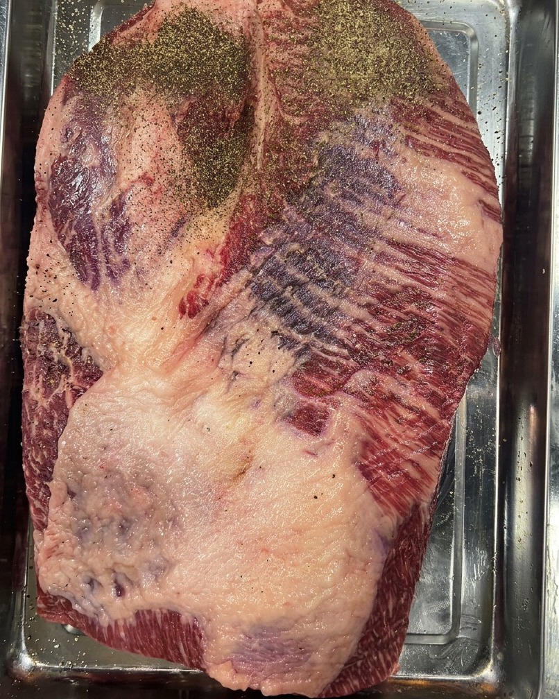 Un brisket de res crudo en una bandeja de acero inoxidable, fuertemente sazonado con una capa de pimienta negra molida.