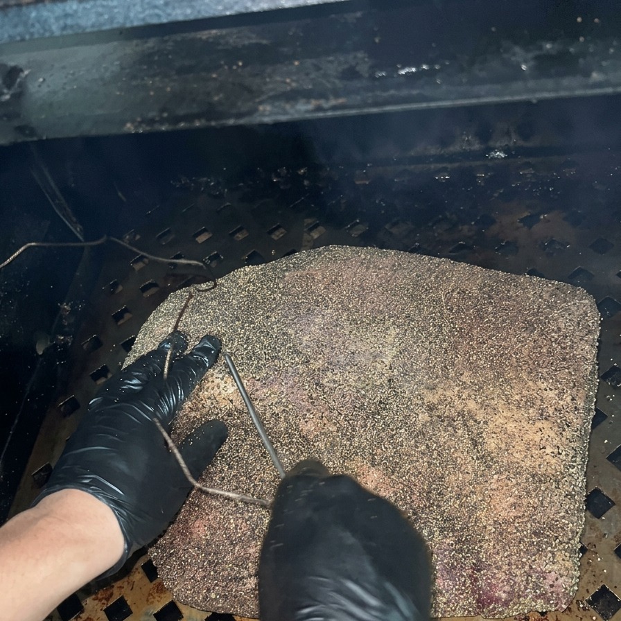 Una persona con guantes negros insertando la sonda de un termómetro metálico en un brisket sazonado dentro de un ahumador oscuro.