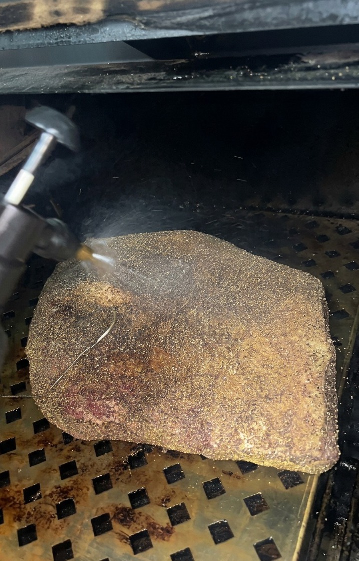 Una fina niebla de vinagre de sidra de manzana siendo rociada sobre un brisket que está desarrollando una corteza oscura dentro del ahumador.