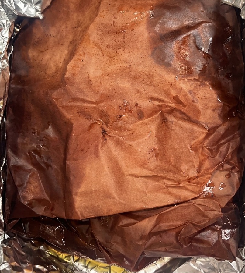 Un gran brisket envuelto en papel de carnicero oscuro y empapado de jugos dentro de un contenedor forrado con aluminio.
