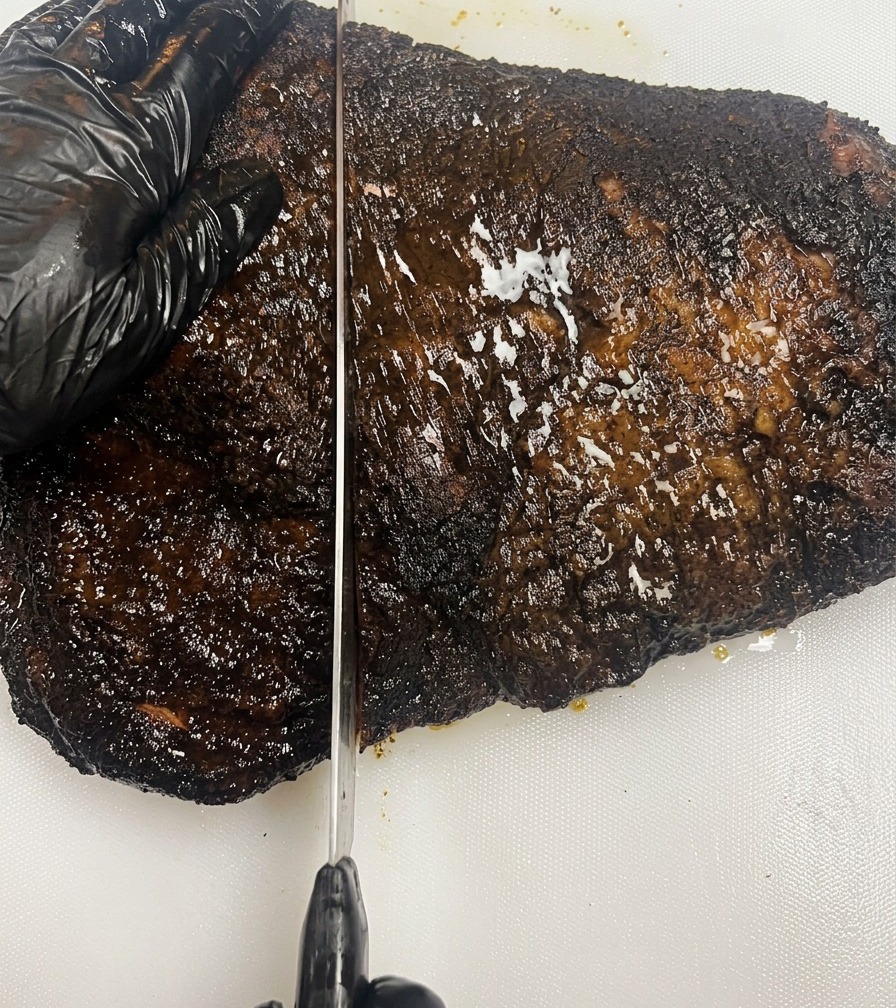 Una mano con guante negro sosteniendo un brisket ahumado de corteza oscura sobre una tabla de cortar blanca mientras un cuchillo largo corta la carne.