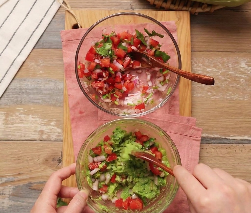Una persona usando una cuchara de madera para incorporar salsa de tomate fresca en un tazón con aguacate machacado.