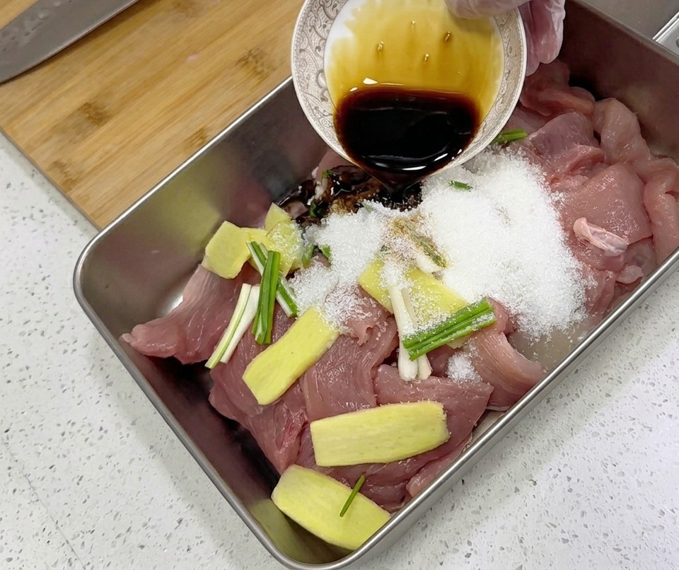 Condimento líquido oscuro vertiéndose desde un pequeño tazón sobre rodajas de cerdo crudo, jengibre y cebolletas en una bandeja de metal.