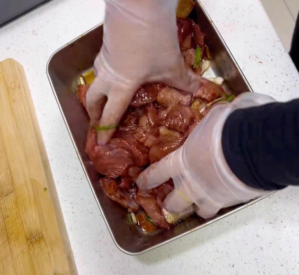 Manos con guantes de plástico mezclando rodajas de carne cruda con una marinada roja oscura en una bandeja de acero.