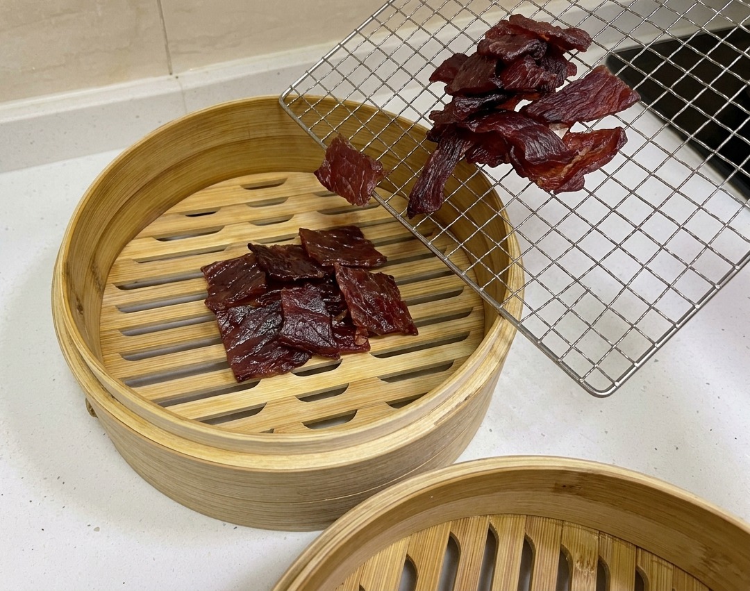 Trozos de cerdo secados al aire de color marrón rojizo oscuro siendo trasladados desde una rejilla a una cesta vaporera circular de bambú.