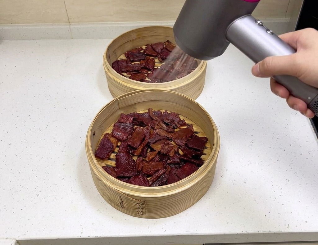Una persona sosteniendo un secador de pelo moderno dirigiendo aire sobre trozos de carne seca marrón oscura en cestas de bambú.