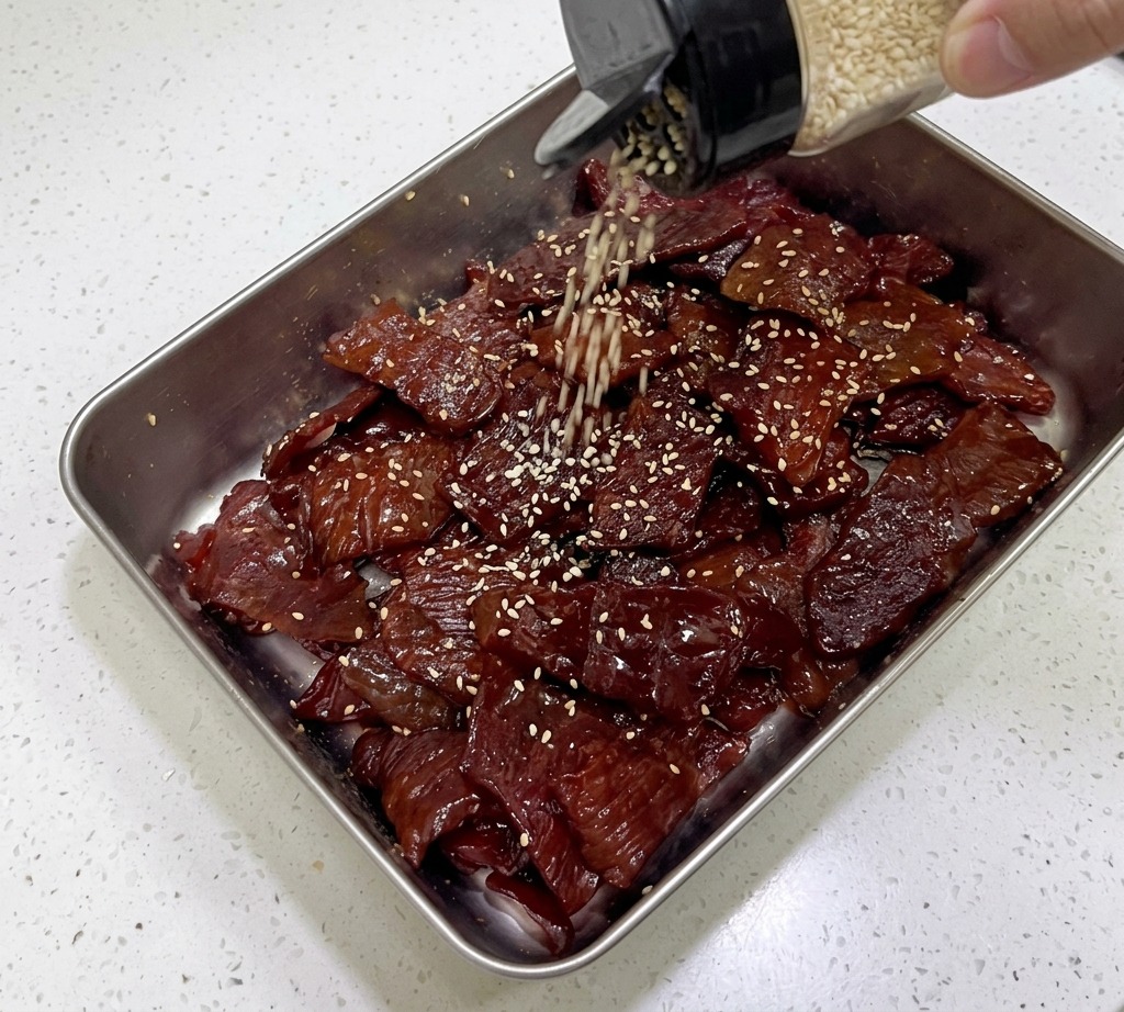 Semillas de sésamo blanco tostadas cayendo de un salero de vidrio sobre tiras de cerdo brillantes y oscuras en una bandeja plateada.