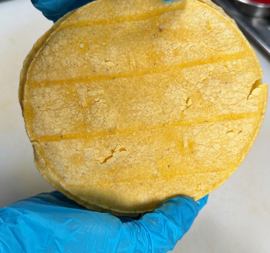 Una persona con guantes azules sosteniendo una pila de tortillas de maíz sobre una superficie de cocina blanca.