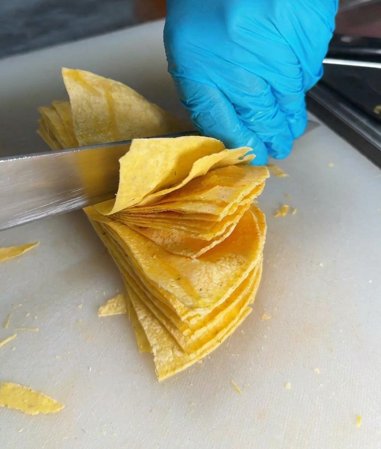 Un cuchillo afilado cortando una pila de tortillas para crear triángulos sobre una tabla blanca.