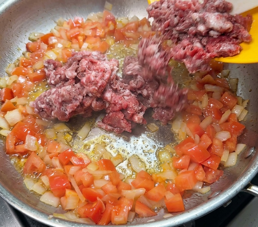 Carne molida cruda siendo añadida a una sartén con sofrito de cebolla y tomate.