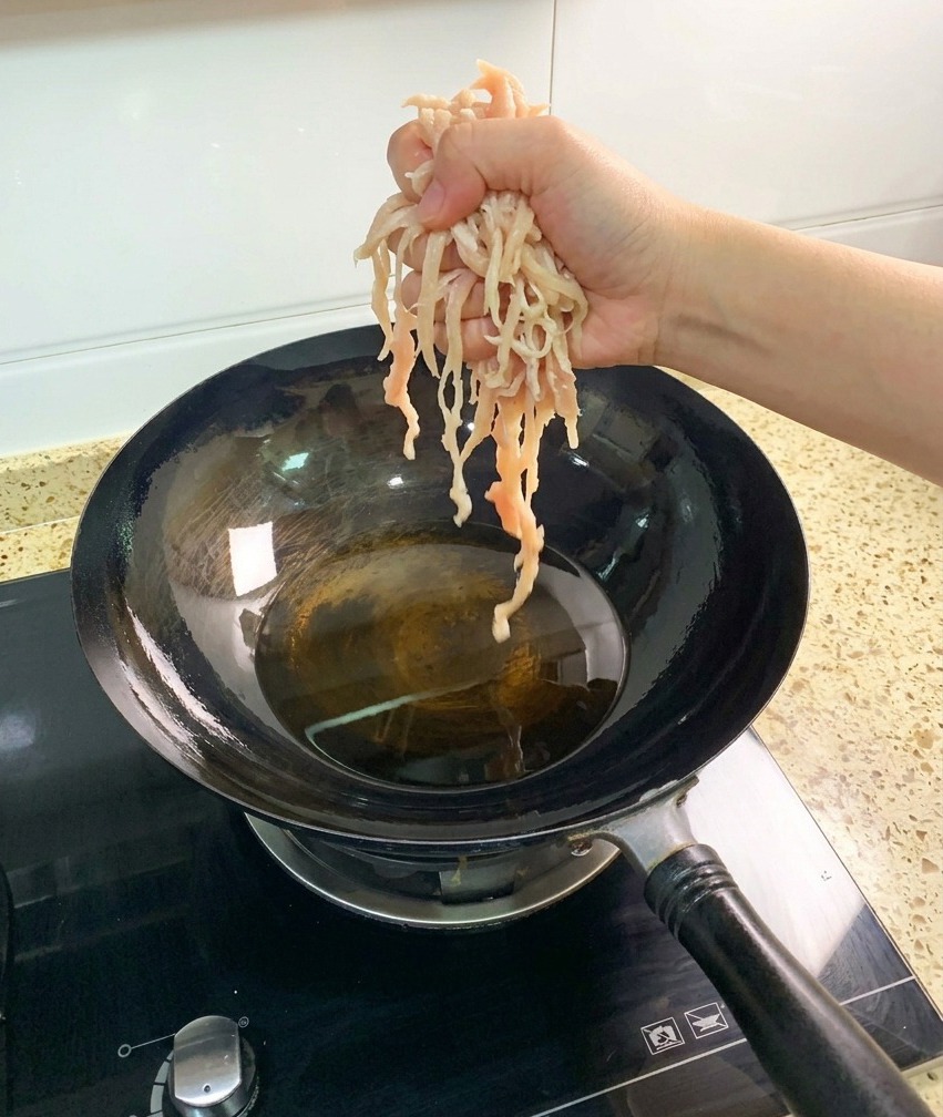 Una mano dejando caer trozos de pechuga de pollo desmechada, cruda y pálida, en un wok oscuro lleno de aceite de cocina caliente y transparente.