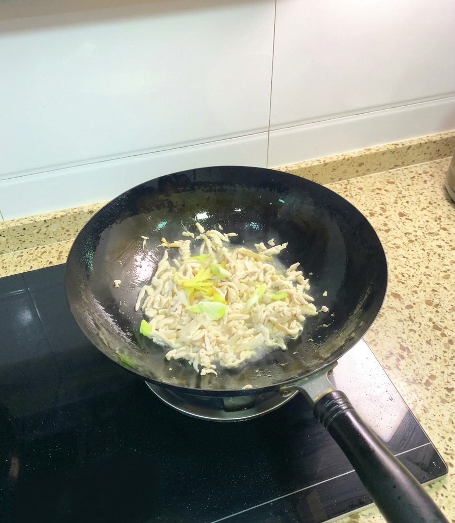 Trozos de pollo opacos y parcialmente cocinados en un wok siendo salteados con trozos de cebolleta fresca y rodajas de jengibre.