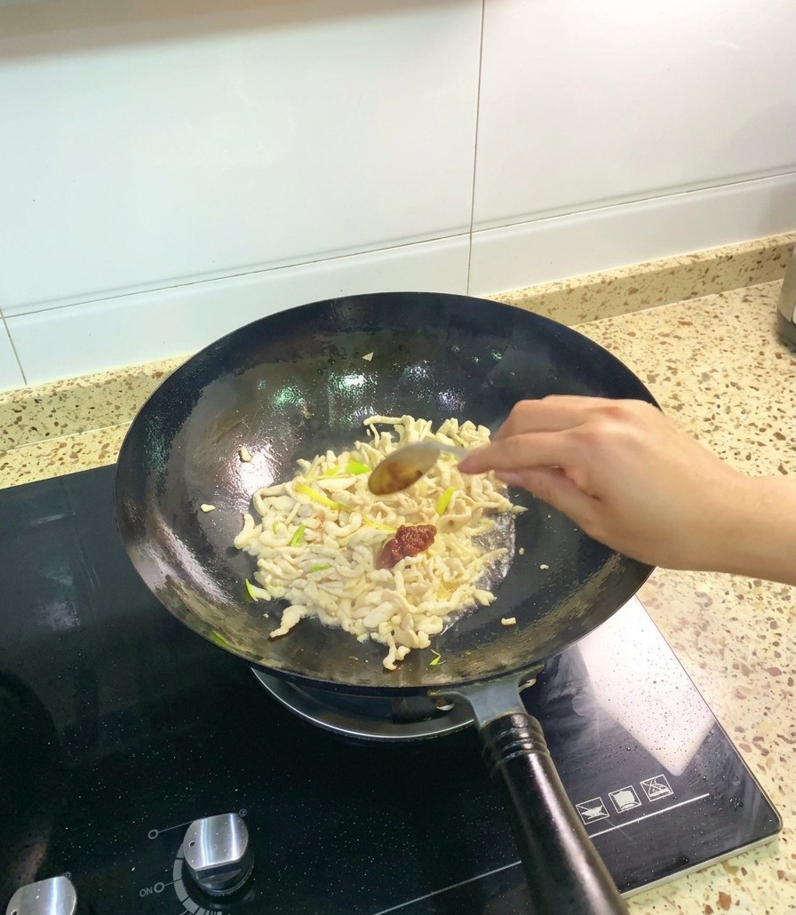 Una cuchara de acero inoxidable agregando una porción de pasta de frijoles de Pixian, espesa y roja, a un wok con pollo salteado y aromáticos.