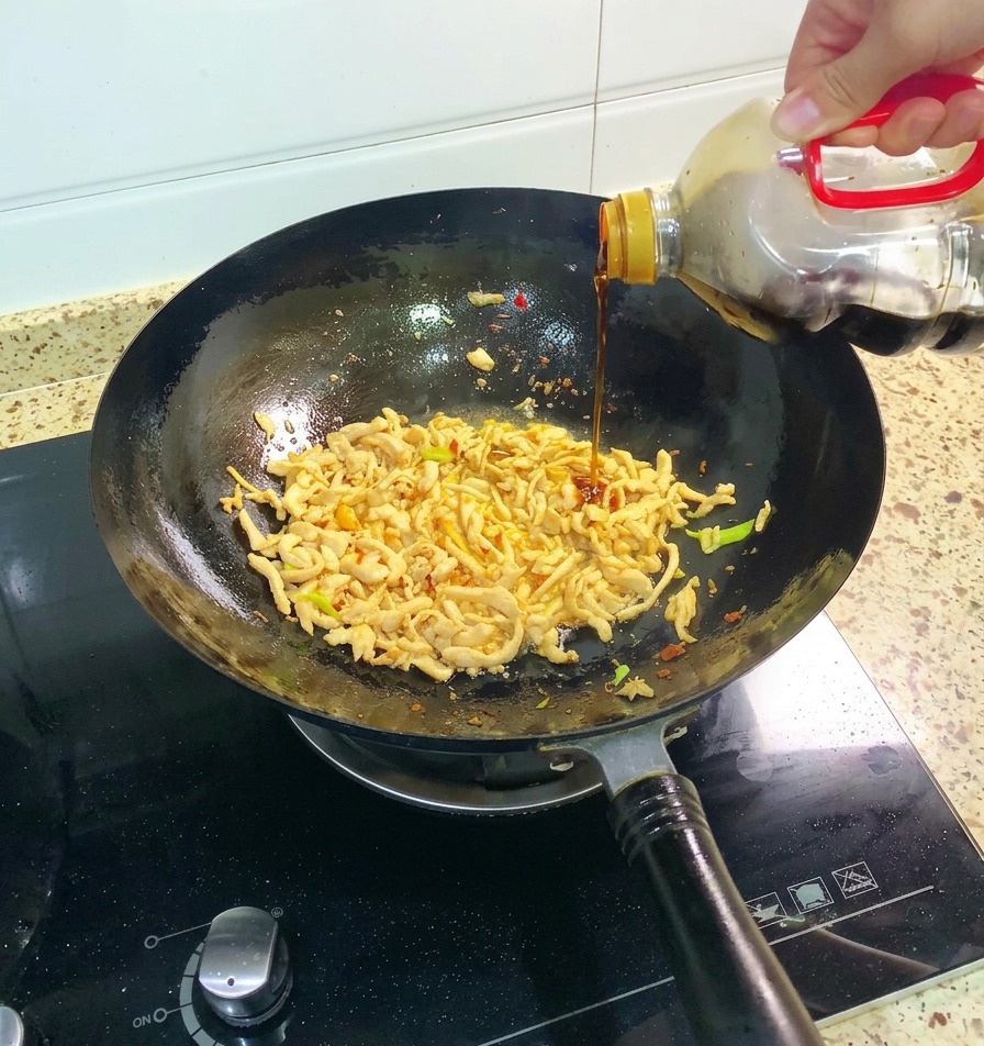 Una mano vertiendo salsa de soja oscura desde una botella grande en un wok que contiene pollo desmechado y aromáticos verdes.