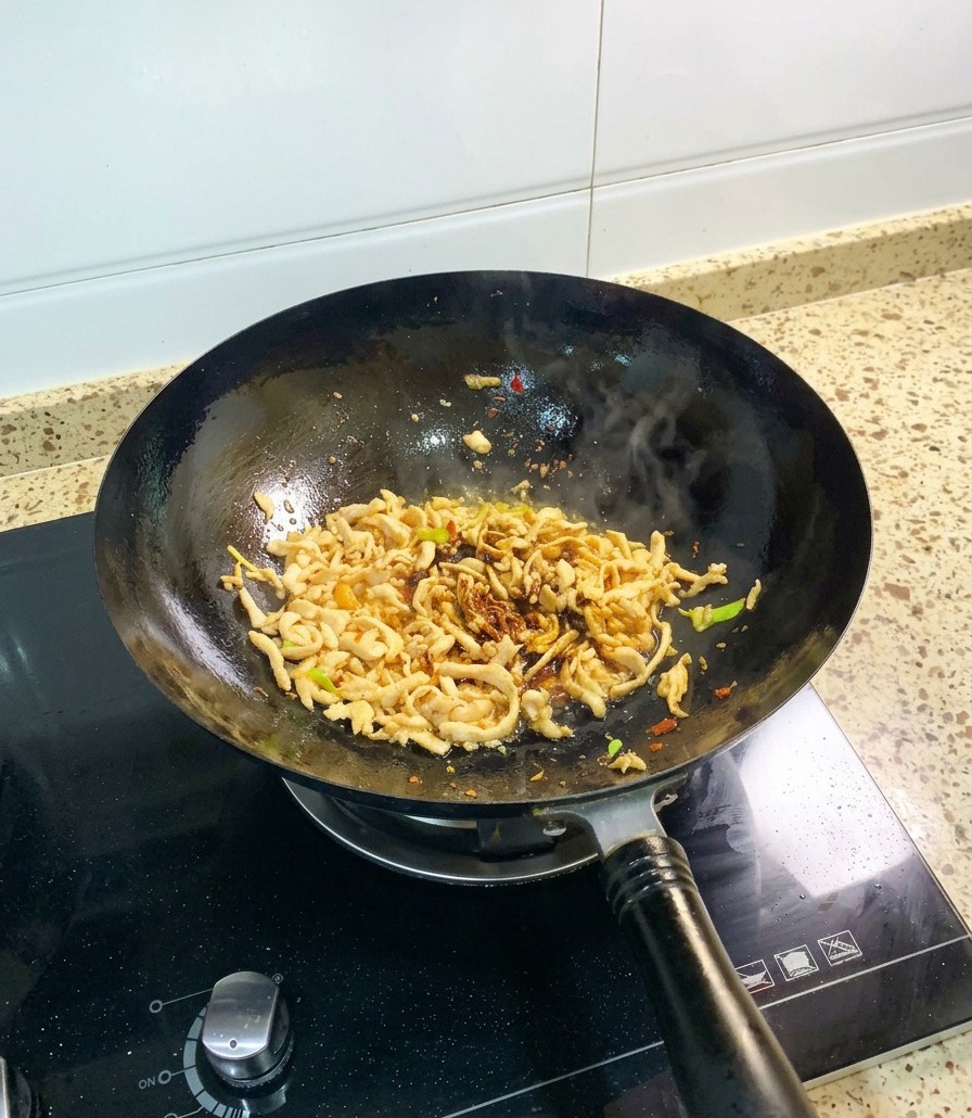 Pollo desmechado en un wok negro siendo salteado con salsa de soja oscura y una adición visible de salsa de ostras espesa.