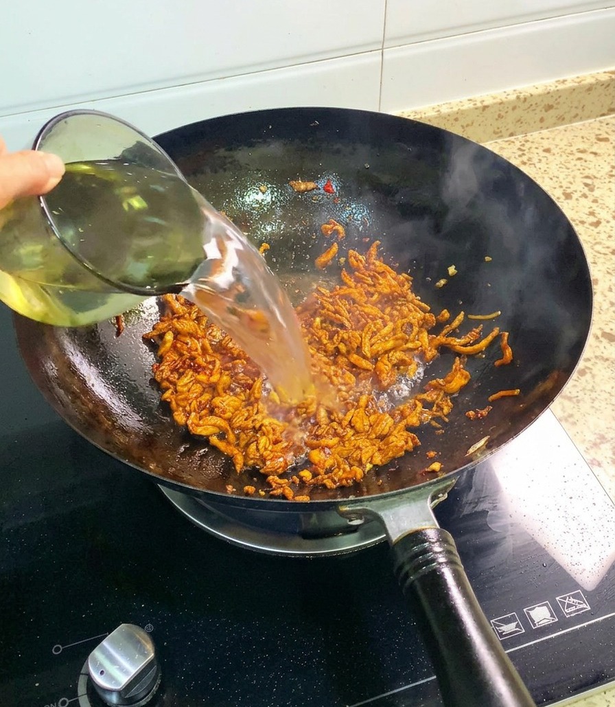 Un chorro de agua limpia siendo vertido desde una taza de vidrio en un wok lleno de pollo desmechado sazonado y salteado.