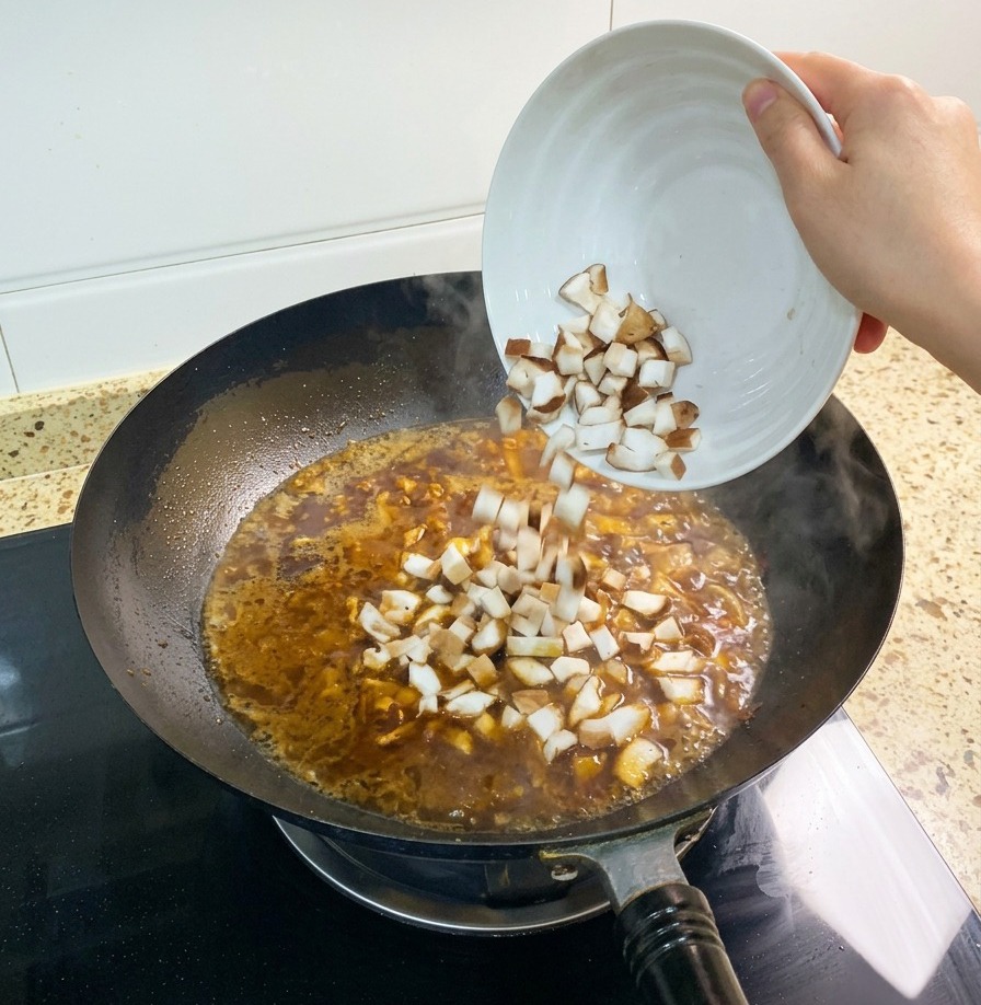 Un puñado de champiñones shiitake picados siendo añadidos al caldo marrón rojizo que hierve a fuego lento en un wok.