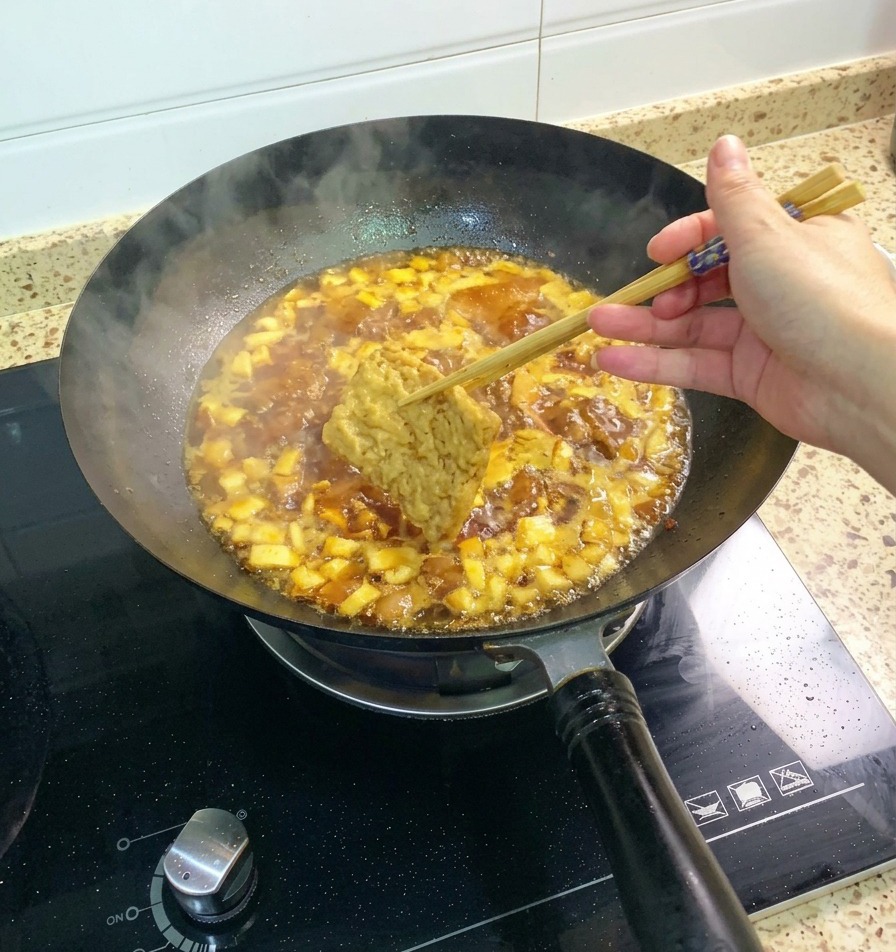 Un trozo de tofu frito sostenido por palillos siendo sumergido en la salsa burbujeante de pollo y champiñones.