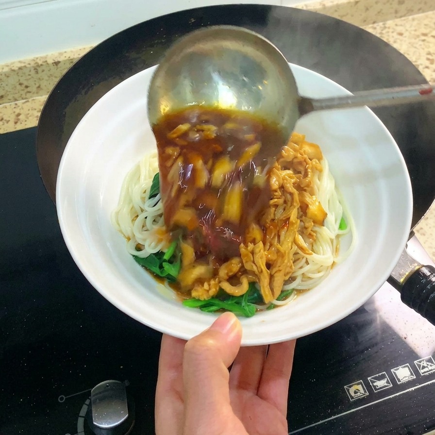 Un cucharón de metal grande vertiendo una salsa oscura y sabrosa de pollo desmechado y champiñones sobre un tazón blanco con fideos.