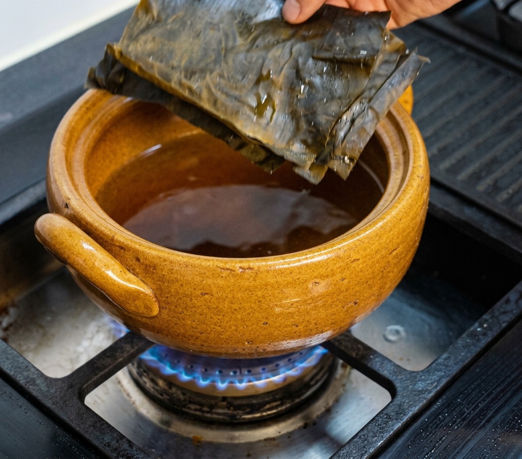 Una mano colocando láminas rectangulares de alga kombu deshidratada de color verde oscuro en una olla de barro marrón sobre el quemador de una estufa de gas.