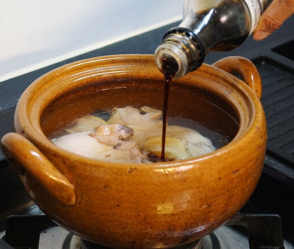 Una mano vertiendo condimento líquido oscuro desde una botella de vidrio en la olla que contiene el pollo y el agua.