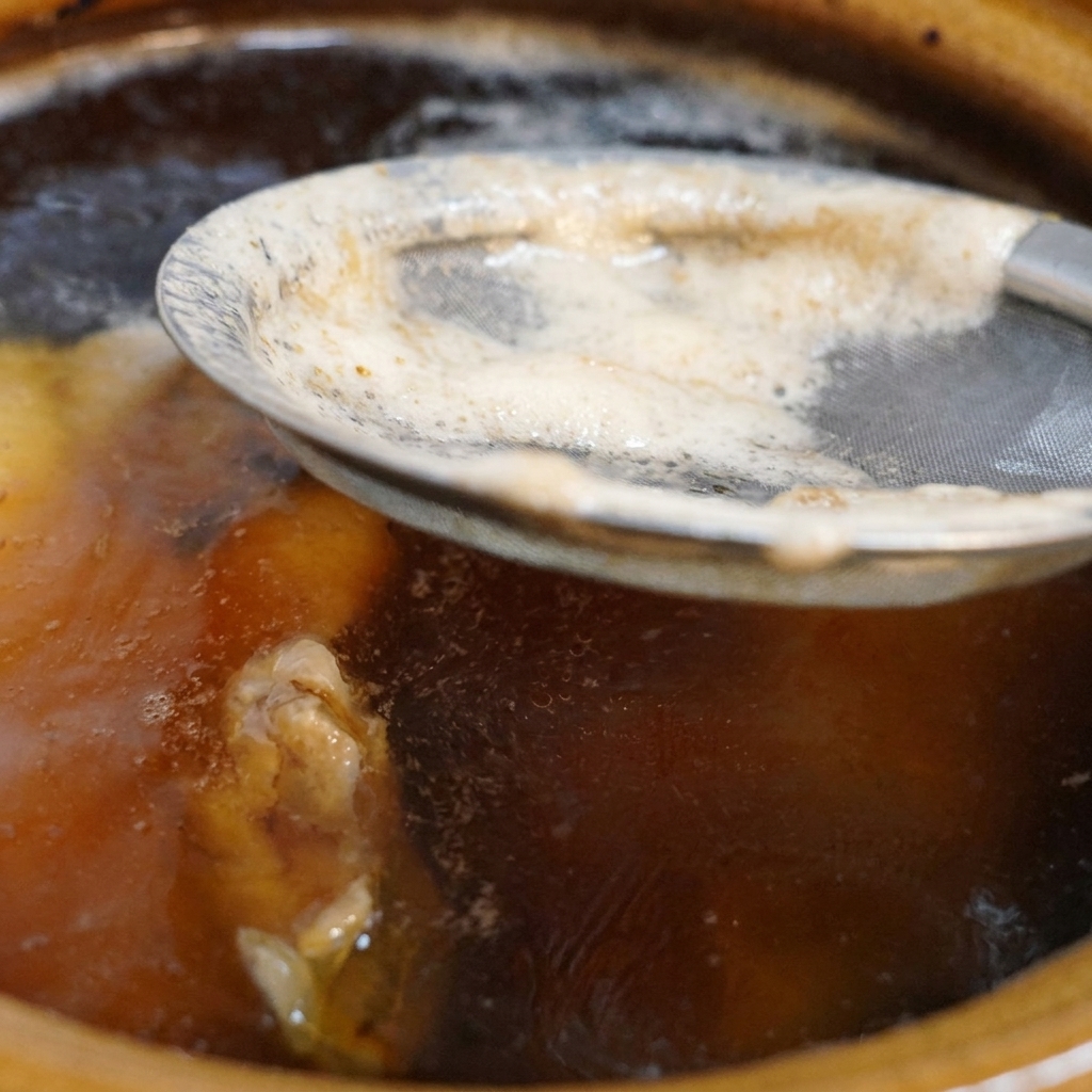 Un colador metálico de malla fina retirando la espuma blanca de la superficie de un caldo de pollo oscuro que hierve en una olla de cerámica.