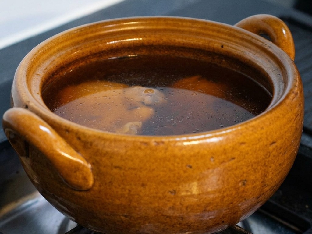 Una olla de cerámica marrón en una estufa de gas llena de un caldo oscuro a base de soja que hierve suavemente, con la tapa parcialmente levantada para revelar el pollo y el kombu en su interior.