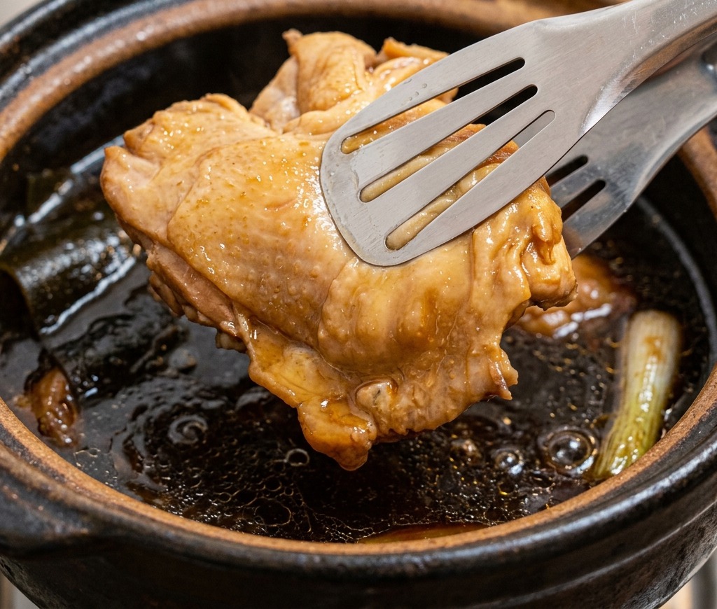 Pinzas de metal levantando un trozo de contramuslo de pollo cocido y dorado de una olla de caldo de pollo oscuro.