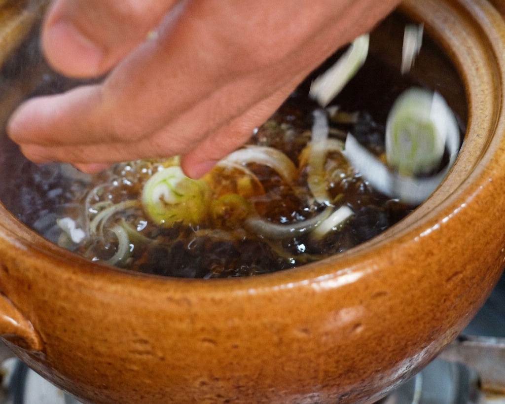 Manos dejando caer un puñado de cebolleta verde picada en una olla con caldo de pollo oscuro hirviendo.