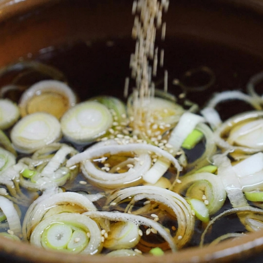 Semillas de sésamo blanco cayendo en un caldo oscuro que hierve a fuego lento con cebolleta picada dentro de una olla de cerámica.