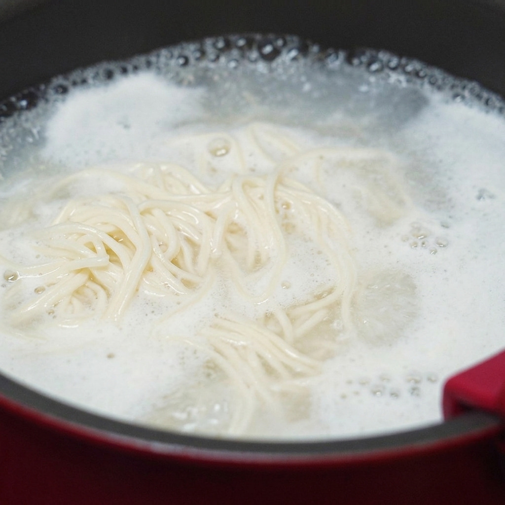 Fideos de trigo blanco girando en una olla de agua hirviendo a borbotones con una densa espuma almidonada que sube hacia los bordes.