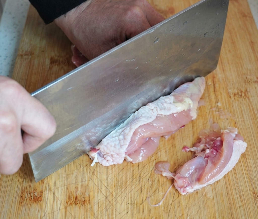 Persona usando un cuchillo de carnicero para deshuesar un muslo de pollo crudo sobre una tabla de cortar de madera.