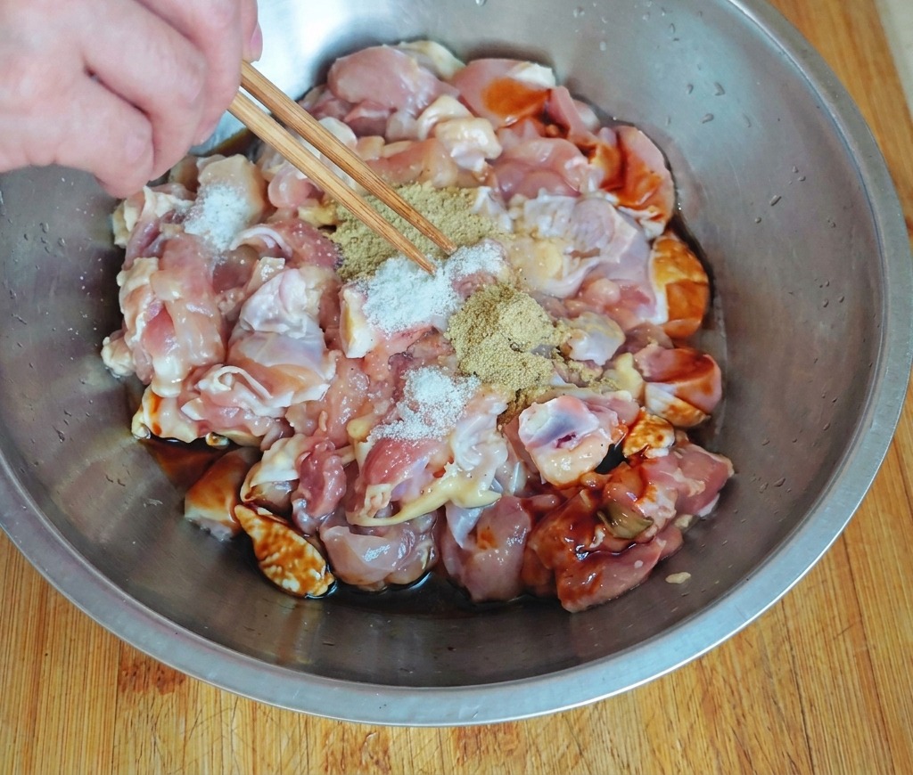 Mano usando palillos para espolvorear condimento blanco sobre trozos de pollo crudo en un bol de acero inoxidable con salsas oscuras.