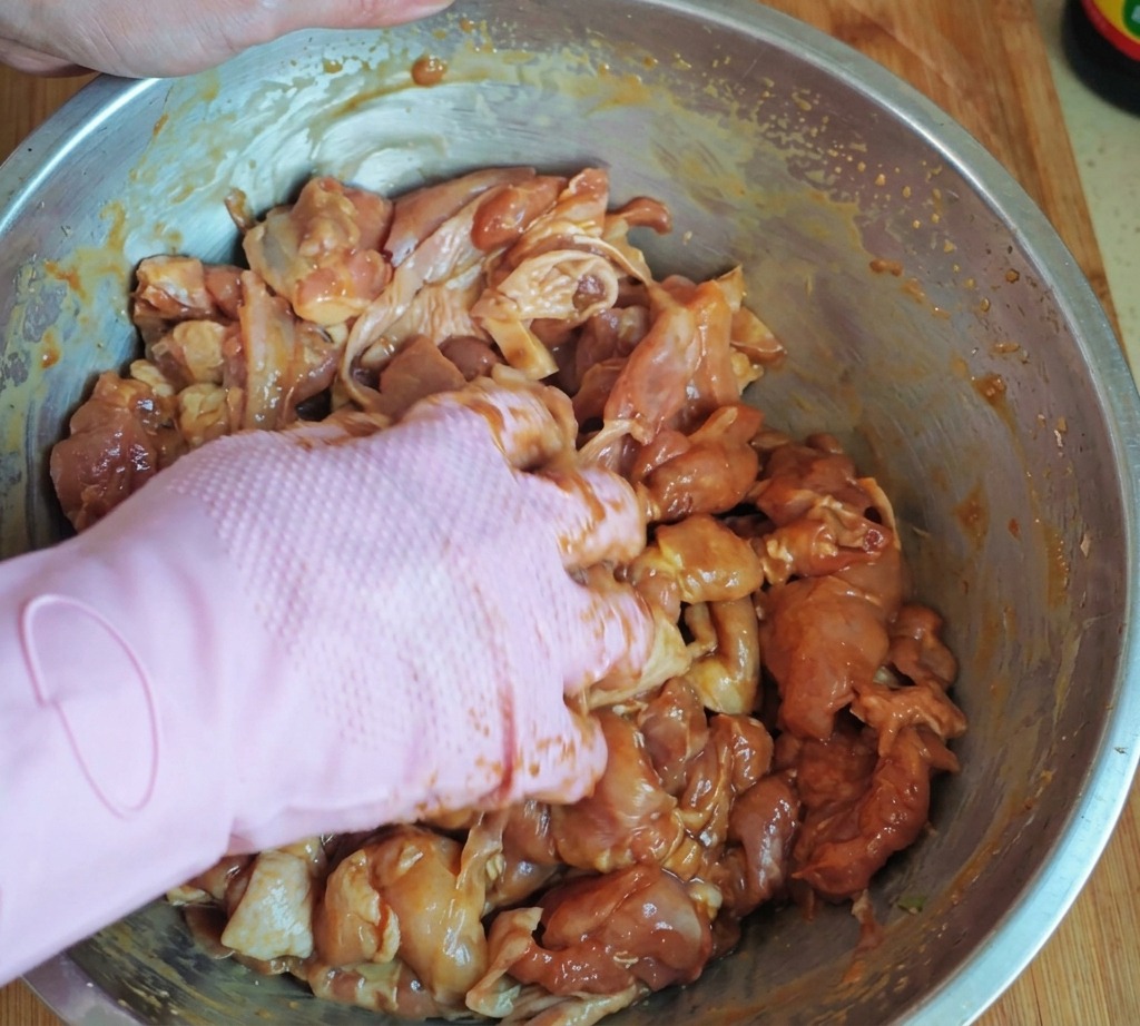 Mano con guante rosa mezclando bien trozos de pollo marinado con almidón y aceite en un bol grande de acero inoxidable.