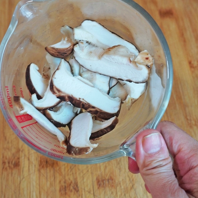 Vista cenital de setas shiitake frescas cortadas en rodajas dentro de una taza medidora de cristal sobre una tabla de madera.