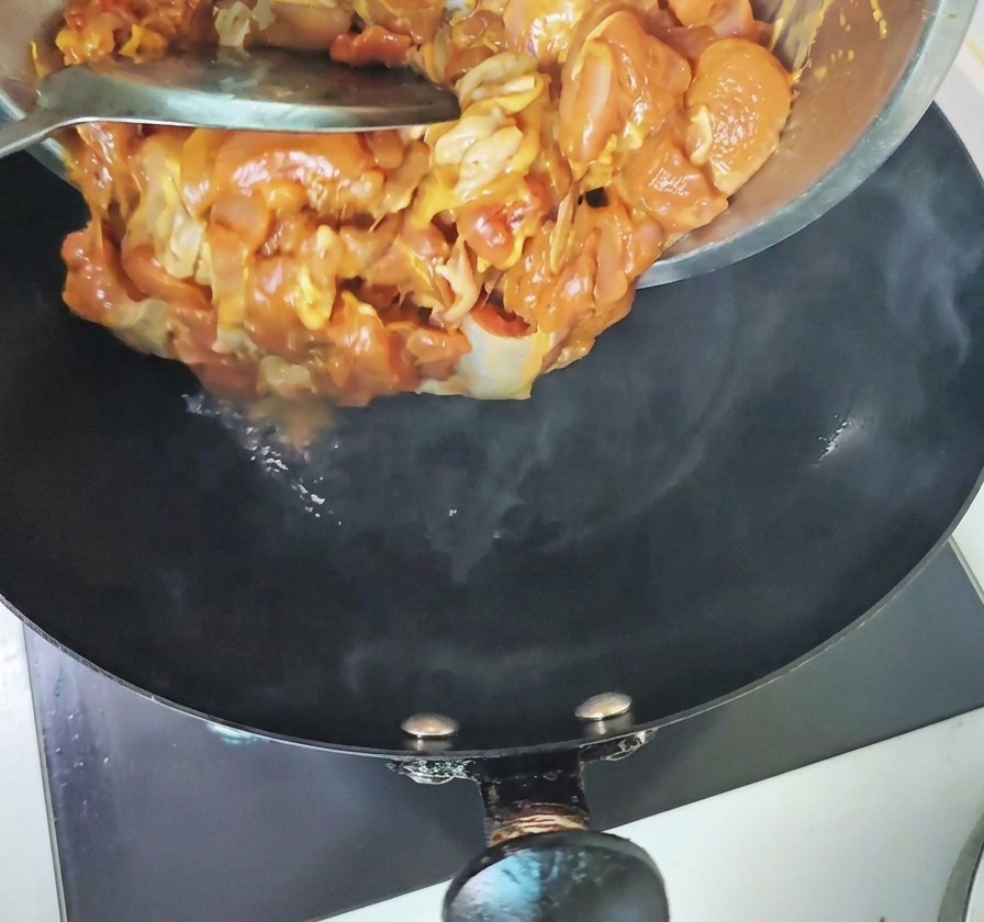 Trozos de pollo marinado vertiéndose desde un bol de metal en un wok negro caliente y humeante.