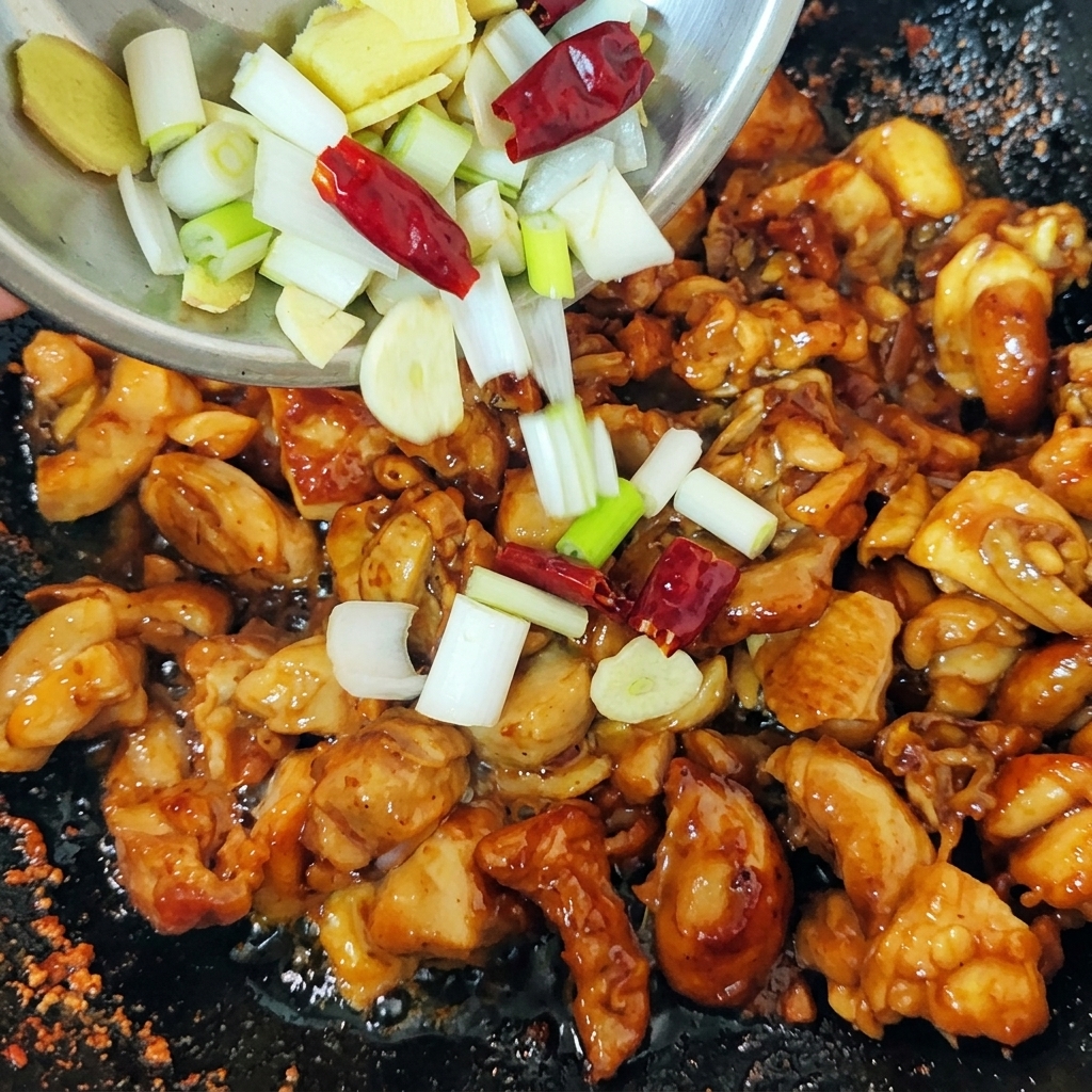 Mezcla de cebolletas verdes picadas, rodajas de jengibre y chiles rojos secos cayendo desde un bol pequeño a un wok con pollo sellado.
